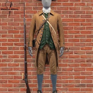 May include: A historical military uniform featuring a tan long coat, a dark green button-up vest, tan breeches, and grey stockings. A white cravat is worn at the neck. A long, dark-colored musket stands vertically to the left of the mannequin.