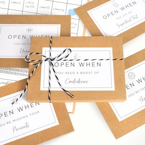 May include: A collection of brown paper cards with white labels and black text. The cards are tied with black and white twine. The text on the cards reads "Open When" followed by various sentiments, such as "You Need a Boost of Confidence."