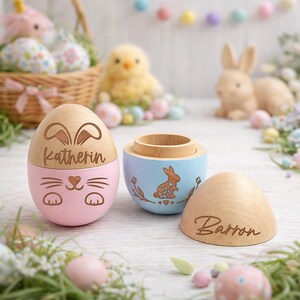 May include: Wooden Easter egg-shaped boxes, one pink with a bunny face and the name "Katherin", and one blue with a bunny illustration. A third wooden egg top is engraved with the name "Barron". The boxes are surrounded by Easter decorations.