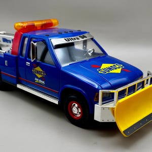 Könnte beinhalten: Ein blaues Spielzeug-Truck mit einem gelben Schneepflug und einem orangefarbenen Lichtbalken. Der Truck hat das "SUNOCO"-Logo und "Ultra 94" aufgedruckt. Die Räder sind rot und der Truck steht auf einer grauen Oberfläche.