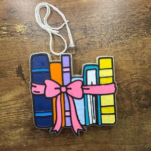 Könnte beinhalten: Ein farbenfroher Ornament in Form eines Bücherstapels, der mit einer rosa Schleife gebunden ist. Die Bücher sind in verschiedenen Farben gehalten, darunter Blau, Orange, Lila und Gelb. Ein weißer Faden ist zum Aufhängen befestigt. Das Ornament befindet sich auf einer Holzoberfläche.