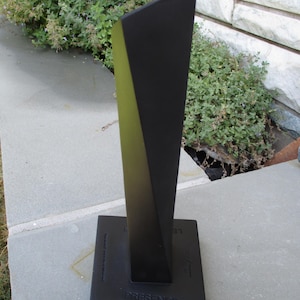 May include: A matte black sculptural object with a tall, tapered, geometric form rising from a rectangular base. The base features the word PRESENCE embossed on the front surface, along with additional faint, partially visible text markings.