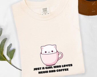 Può includere: T-shirt color avorio con grafica di un gatto bianco in una tazza da caffè rosa. Il testo "JUST A GIRL WHO LOVES MEOW AND COFFEE" è stampato sotto. Marchio Comfort Colors.