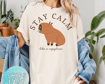 May include: Ivory-colored t-shirt with the text "STAY CALM" above a cartoon capybara illustration. The phrase "like a capybara" is printed below the image. The shirt has a relaxed fit.
