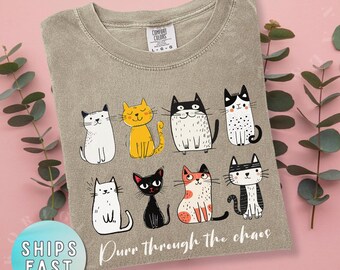 May include: Khaki-colored t-shirt featuring a collection of cartoon cats in various poses and colors. The phrase "Purr through the chaos" is printed below the cats. The shirt has a crew neck and short sleeves.
