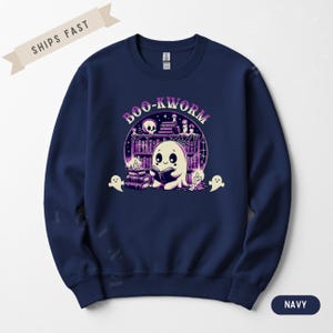 Puede incluir: Sudadera azul marino con un fantasma de dibujos animados leyendo un libro frente a una estantería. El diseño incluye el texto "BOO-KWORMS" en morado, además de fantasmas, velas y una calavera. Prenda cómoda e informal.