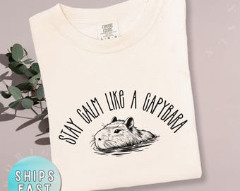 May include: Ivory-colored t-shirt with the text "Stay Calm Like a Capybara" in a curved font above a black and white illustration of a capybara in water. The shirt is a Comfort Colors brand.