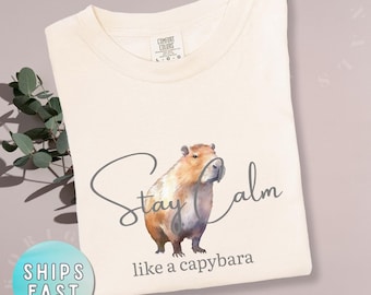 May include: Ivory-colored t-shirt featuring a watercolor illustration of a capybara. The shirt has the text "Stay Calm like a capybara" in a script font. The shirt is made by Comfort Colors.
