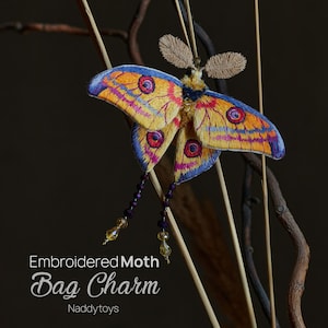 Peut inclure: Charm de sac en forme de papillon de nuit brodé aux ailes jaunes, bleues, violettes et oranges. Le charm est orné de brins perlés. Le texte "Embroidered Moth Bag Charm" est visible.