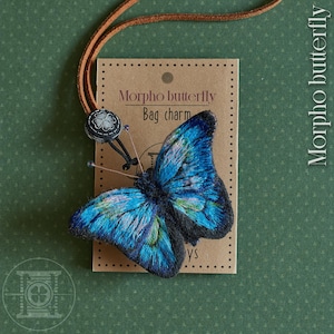 May include: A detailed blue and black butterfly bag charm with a brown leather cord. The charm is attached to a brown tag that reads "Morpho butterfly Bag charm". The butterfly has intricate details and a four-leaf clover charm.