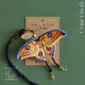 May include: A Comet moth charm with embroidered wings in yellow, orange, purple, and blue. The charm has a beaded strand and a tag that reads "Mother the World charm". The charm is on a green background.