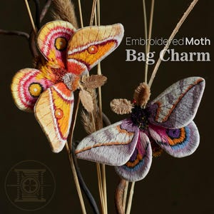 Op de afbeelding: Twee geborduurde motten-tashangers. De ene mot heeft oranje, gele en roze vleugels. De andere mot heeft paarse, grijze en blauwe vleugels. De tekst "Embroidered Moth Bag Charm" is zichtbaar.