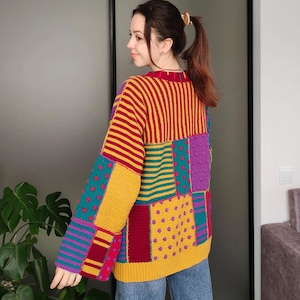 May include: A vibrant, patchwork sweater with a variety of patterns and colours, including stripes, polka dots, and solid blocks. The sweater has a yellow base with red, teal, purple, and burgundy accents. The sleeves are long, and the neckline is a deep red.