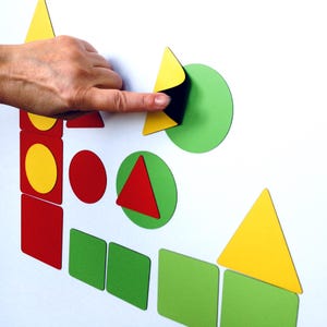 May include: A selection of brightly coloured geometric shapes, including triangles, circles, and squares, in red, yellow, and green. A hand is shown interacting with the shapes, which appear to be magnetic or adhesive. The shapes are arranged on a white surface.