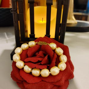 May include: A pearl bracelet with a gold clasp rests on a red rose. The pearls are off-white and have a smooth, rounded shape. The bracelet is the focal point, with a candle and a decorative lantern in the background.
