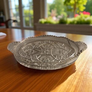May include: A clear, decorative glass serving dish with a detailed floral and geometric pattern. The dish has a scalloped edge and two small handles. The dish is sitting on a wooden table.