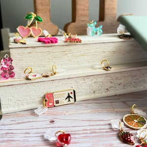 May include: A collection of colorful phone charms featuring various designs, including cherries, hearts, a gummy bear, and a travel ticket with the text "Adventure Awaits". The charms are made of metal and acrylic, with gold-colored accents.