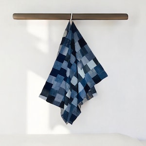 Può includere: Un asciugamano a quadri blu e bianchi è appeso a una barra di legno su un muro bianco. L'asciugamano è composto da quadrati di varie tonalità di blu. Un gancio bianco collega l'asciugamano alla barra.