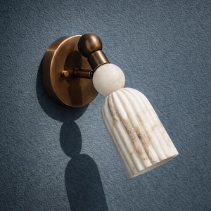 May include: A wall-mounted light fitting with a brass finish and a white, vertically ribbed shade. The fitting has a spherical accent and is mounted on a blue wall. The shade has a marble-like appearance.