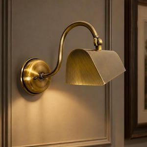 May include: A wall-mounted lamp with an antique brass finish, featuring a circular base, a curved arm, and a rectangular, downward-angled shade. The fixture emits a soft, warm glow from beneath the metal hood, highlighting the metallic texture.