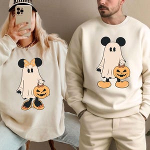 Könnte beinhalten: Cremefarbene Sweatshirts mit einem Cartoon-Geist mit Mickey Mouse-Ohren, der einen Kürbis-Laterne hält. Der Geist hat schwarze Ohren, ein weißes Laken und orangefarbene Schuhe. Ein Sweatshirt hat eine Schleife. Halloween-Themenbekleidung.