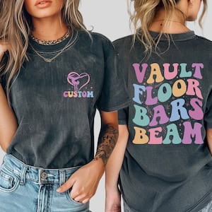 May include: Dark gray t-shirts with a gymnastics theme. The front of the shirt has a heart-shaped design with a gymnast and the word "CUSTOM". The back of the shirt has the words "VAULT FLOOR BARS BEAM" in colorful, retro-style lettering.