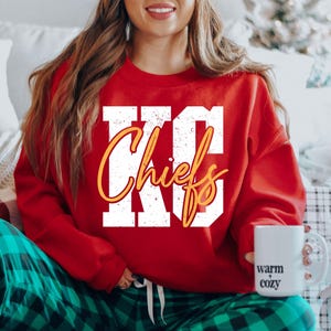Könnte beinhalten: Ein rotes Sweatshirt mit dem Text "KC Chiefs" in weißen Blockbuchstaben mit gelbem Schriftzug. Die Person trägt eine grüne karierte Pyjamahose und hält einen weißen Becher mit der Aufschrift "warm cozy".