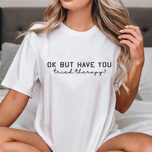 May include: White t-shirt with the text "OK BUT HAVE YOU tried therapy?" in black. The shirt has a crew neck and short sleeves. The image shows the shirt on a person sitting on a bed.