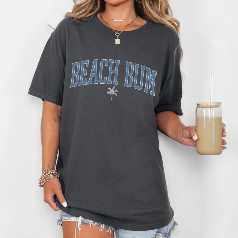 a woman holding a drink and wearing a beach bum t - shirt