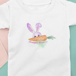 May include: White toddler t-shirt featuring a watercolour illustration of a purple bunny holding a carrot in its mouth. The text "Happy Easter" is printed below the bunny.