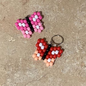 Puede incluir: Dos dijes con forma de mariposa hechos con peque&ntilde;as cuentas de pl&aacute;stico. Uno presenta cuentas rosas, blancas y negras, mientras que el otro muestra cuentas rojas, blancas, negras y color melocot&oacute;n. Cada mariposa incluye una peque&ntilde;a argolla.