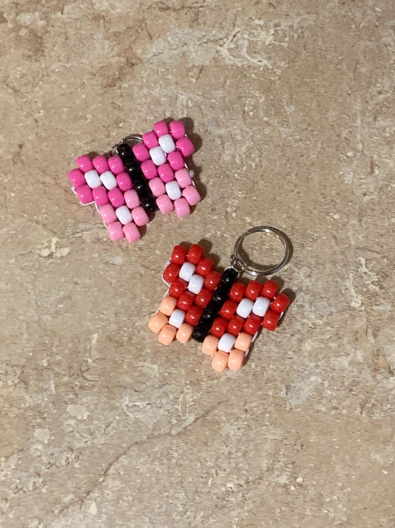 Puede incluir: Dos dijes con forma de mariposa hechos con peque&ntilde;as cuentas de pl&aacute;stico. Uno presenta cuentas rosas, blancas y negras, mientras que el otro muestra cuentas rojas, blancas, negras y color melocot&oacute;n. Cada mariposa incluye una peque&ntilde;a argolla.