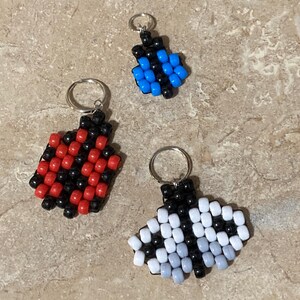 Puede incluir: Tres llaveros hechos de peque&ntilde;as cuentas de pl&aacute;stico. Un llavero presenta un patr&oacute;n de cuadros rojo y negro, otro es un dise&ntilde;o m&aacute;s peque&ntilde;o azul y negro, y el tercero es un patr&oacute;n blanco, gris y negro, cada uno con una anilla de metal.