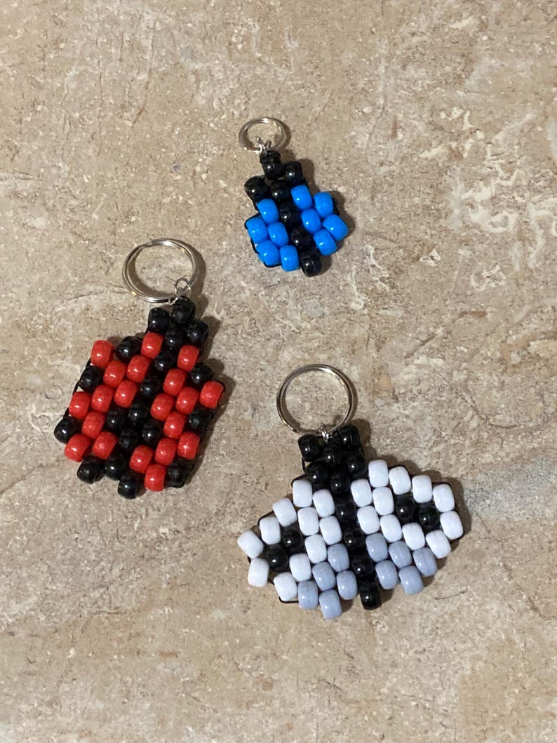 Puede incluir: Tres llaveros hechos de peque&ntilde;as cuentas de pl&aacute;stico. Un llavero presenta un patr&oacute;n de cuadros rojo y negro, otro es un dise&ntilde;o m&aacute;s peque&ntilde;o azul y negro, y el tercero es un patr&oacute;n blanco, gris y negro, cada uno con una anilla de metal.