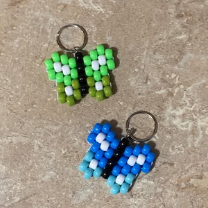 Puede incluir: Dos llaveros con forma de mariposa hechos de cuentas de pl&aacute;stico. Uno tiene cuentas de color verde lima, verde oliva, blanco y negro. El segundo tiene cuentas de color azul claro, azul oscuro, blanco y negro. Cada uno tiene una anilla de metal.