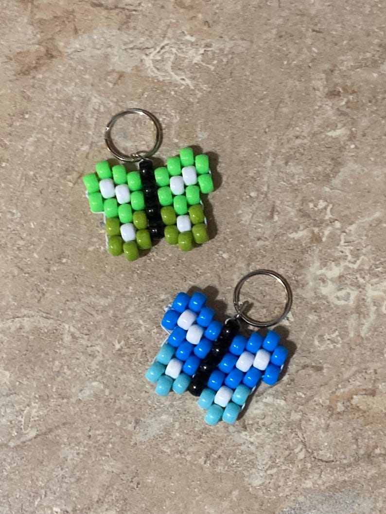 Puede incluir: Dos llaveros con forma de mariposa hechos de cuentas de pl&aacute;stico. Uno tiene cuentas de color verde lima, verde oliva, blanco y negro. El segundo tiene cuentas de color azul claro, azul oscuro, blanco y negro. Cada uno tiene una anilla de metal.