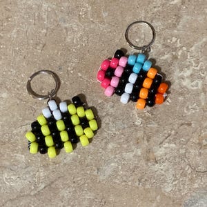 Puede incluir: Dos llaveros hechos con cuentas de pl&aacute;stico. Uno presenta un patr&oacute;n de abeja con cuentas amarillas, negras y blancas. El segundo llavero muestra un patr&oacute;n multicolor con cuentas rosas, azul claro, blancas, negras y naranjas.