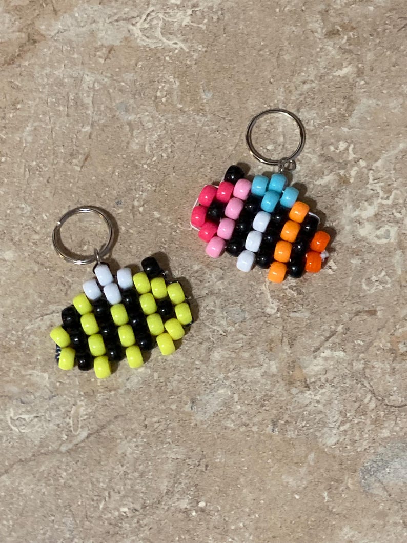 Puede incluir: Dos llaveros hechos con cuentas de pl&aacute;stico. Uno presenta un patr&oacute;n de abeja con cuentas amarillas, negras y blancas. El segundo llavero muestra un patr&oacute;n multicolor con cuentas rosas, azul claro, blancas, negras y naranjas.