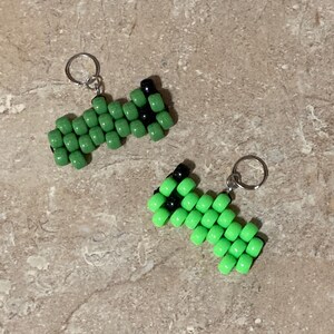 Puede incluir: Dos peque&ntilde;os dijes hechos de cuentas de pl&aacute;stico de color verde lima y negro dispuestas en un patr&oacute;n geom&eacute;trico. Cada dije cuenta con una argolla de metal plateado unida a un extremo para facilitar su uso en llaveros o accesorios.