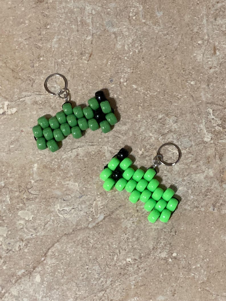 Puede incluir: Dos peque&ntilde;os dijes hechos de cuentas de pl&aacute;stico de color verde lima y negro dispuestas en un patr&oacute;n geom&eacute;trico. Cada dije cuenta con una argolla de metal plateado unida a un extremo para facilitar su uso en llaveros o accesorios.