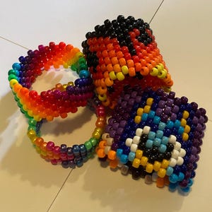 May include: A collection of colorful beaded accessories. Includes a rainbow-colored bracelet, a black, orange, and yellow cuff, and a purple, blue, and white beaded square with a design. The items are made of small, round beads.