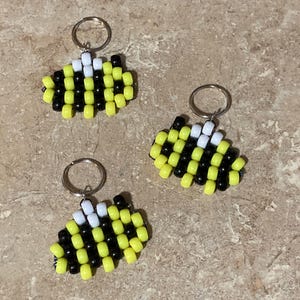 Puede incluir: Tres llaveros con forma de abeja hechos de peque&ntilde;as cuentas de pl&aacute;stico cil&iacute;ndricas en amarillo, negro y blanco. Cada llavero cuenta con una anilla met&aacute;lica en la parte superior, con las cuentas dispuestas en un patr&oacute;n de rayas.