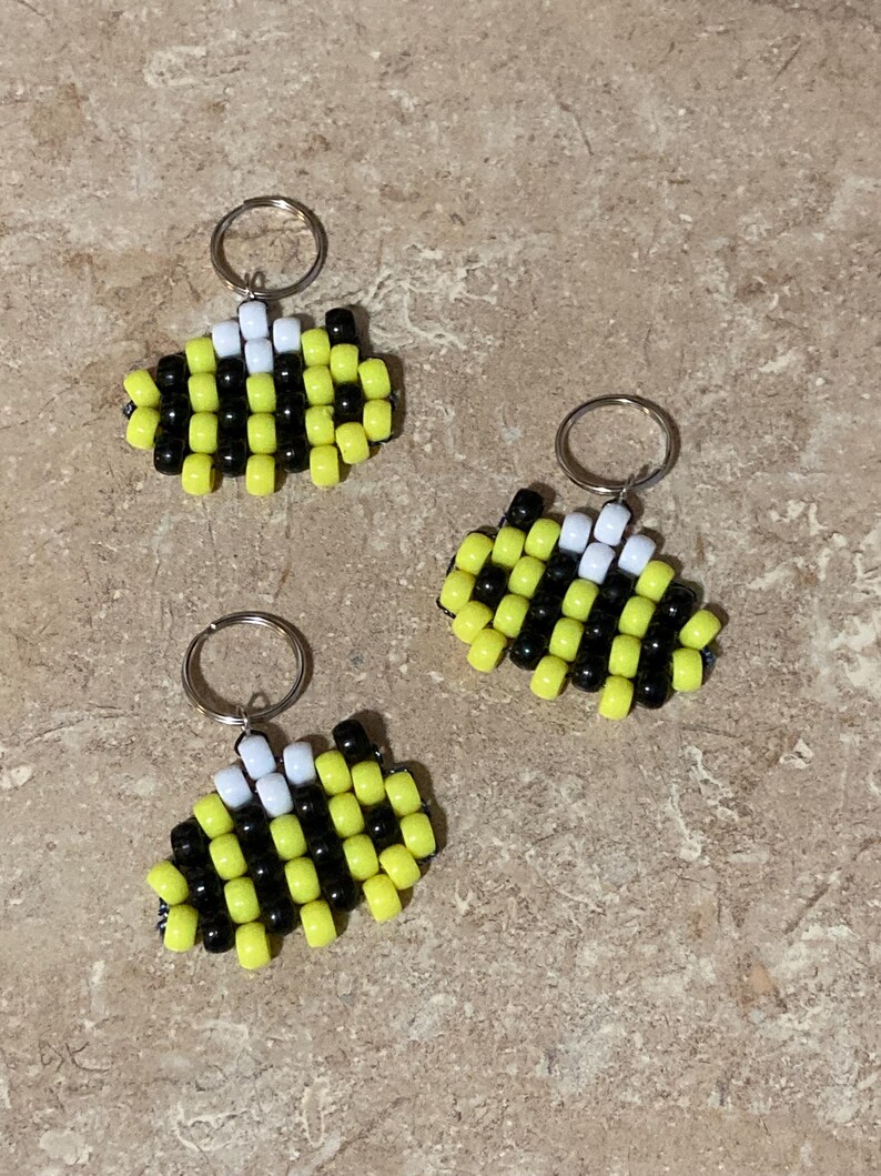 Puede incluir: Tres llaveros con forma de abeja hechos de peque&ntilde;as cuentas de pl&aacute;stico cil&iacute;ndricas en amarillo, negro y blanco. Cada llavero cuenta con una anilla met&aacute;lica en la parte superior, con las cuentas dispuestas en un patr&oacute;n de rayas.