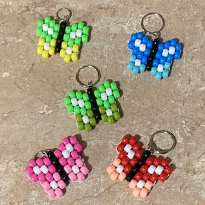 Puede incluir: Cinco llaveros con forma de mariposa hechos de cuentas de pl&aacute;stico de colores. Cada mariposa presenta un cuerpo central de cuentas negras, cuentas blancas de acento y alas en distintas combinaciones de colores verde, amarillo, azul, rosa y rojo.