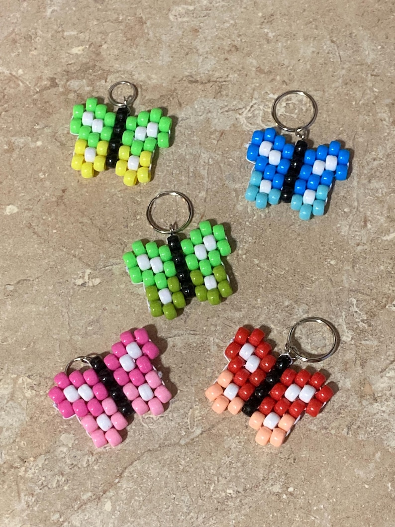 Puede incluir: Cinco llaveros con forma de mariposa hechos de cuentas de pl&aacute;stico de colores. Cada mariposa presenta un cuerpo central de cuentas negras, cuentas blancas de acento y alas en distintas combinaciones de colores verde, amarillo, azul, rosa y rojo.