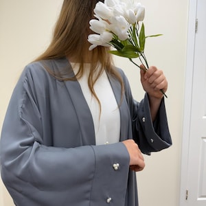 May include: A gray abaya with pearl button details on the sleeves. The abaya is worn over a white garment. A bouquet of white tulips is held in front of the face.