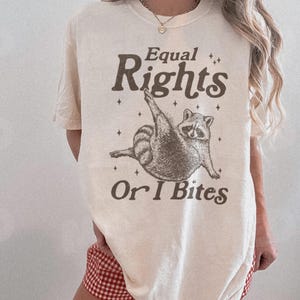 Pode incluir: Camiseta cor marfim com a frase "Equal Rights Or I Bites" e um desenho de guaxinim. O guaxinim é retratado em uma pose divertida. A camiseta tem um corte folgado.