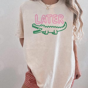 May include: Ivory-colored t-shirt with the word "LATER" in pink above a green alligator graphic. The shirt is a casual style.
