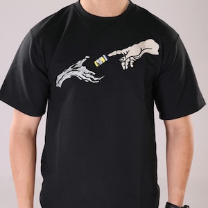 Puede incluir: Camiseta negra con una imagen bordada en blanco de dos manos que se acercan, una sosteniendo una botella amarilla y azul.