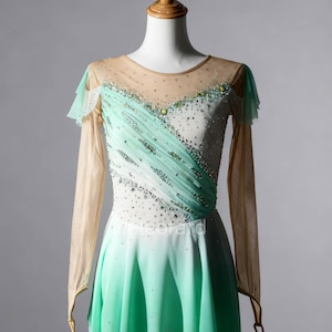 May include: A figure skating dress on a mannequin. The dress features a white bodice with a diagonal mint green overlay, embellished with rhinestones. It has long, sheer sleeves and a mint green skirt. The neckline is sheer and adorned with crystals.